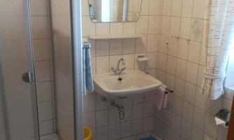 A small bathroom with a shower, washbasin, and a blue doormat with a star motif. The walls are tiled, and there is a window with a white curtain.