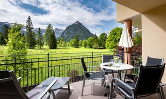 A cozy balcony with seating and a table. In the background, green meadows and majestic mountains can be seen.