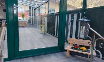 A bright, modern conservatory with a glass wall. In front of the door is a small wooden bench with flowers.