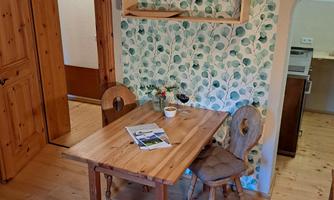 A cozy dining area with a wooden table and two chairs. The walls are decorated with leafy wallpaper, and on the table are a magazine and a small flower vase.