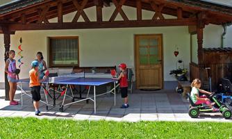 A group of children is playing table tennis on a covered terrace. Another child is riding a toy car in the grass.