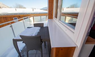 A balcony with a table and chairs is partially covered with snow. The view shows bright, clear weather and a surrounding area with mountains.