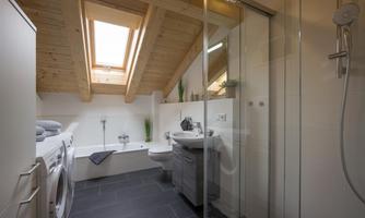 A modern bathroom with sloping walls and a wooden ceiling. It includes a shower, a bathtub, and a washing machine.