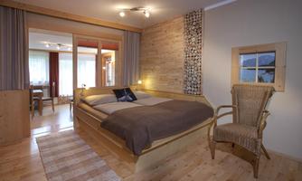 A cozy bedroom with a wooden bed and beige bedding. A wicker chair next to the bed and large windows provide plenty of light.