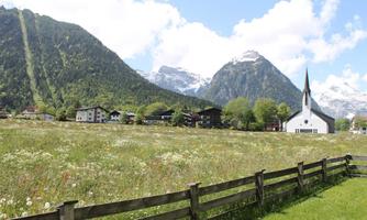 A picturesque landscape with mountains and a church in the foreground. The meadow is blooming and the weather is clear and sunny.
