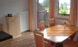 A bright living room with a dining table and chairs. Large windows provide a view outside.
