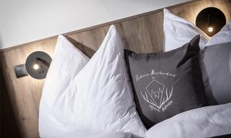 A cozy bed with large white pillows and a decorative pillow in gray. The walls are made of wood, and there are modern wall lamps.