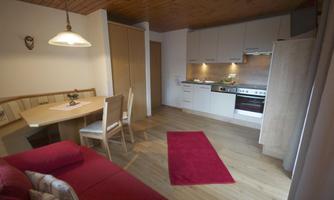 A modern kitchen with a dining table and chairs in a cozy room. A red carpet lies on the floor and a sofa provides comfort.