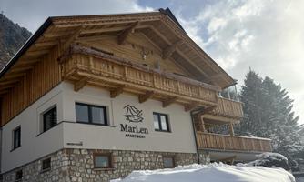 A cozy apartment building with wooden cladding in a snowy mountain landscape. The logo "MarLen Apartment" is visible on the building.