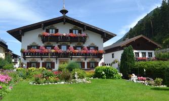 A traditional alpine house with beautiful balconies adorned with flowers. The well-maintained garden with colorful flowers and green areas surrounds the building.