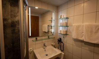 A modern bathroom with a sink, a mirror, and illuminated shelves. Fresh towels hang on the wall, and the tiles are bright and clean.