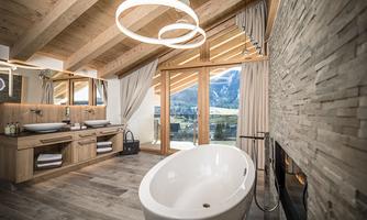 A modern bathroom with a wooden ceiling and large windows. The freestanding bathtub offers a view of the mountains.