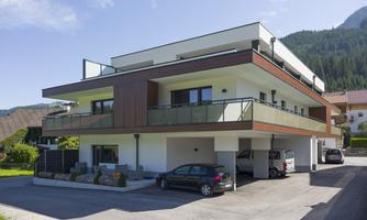 A modern residential building with several floors and large windows. It is located in a quiet environment with green spaces and mountains in the background.