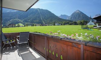 A balcony with two chairs and a beautiful view of green meadows and majestic mountains. The sky is clear and sunny.