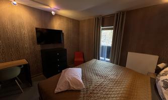 A cozy bedroom with a large bed, a television, and a desk. The room has warm tones and a view of the window.