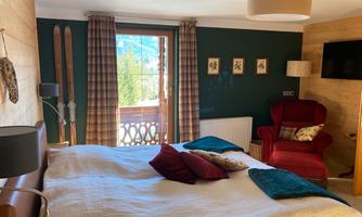 A cozy bedroom with a large bed, a red armchair, and skis on the wall. Large windows offer a view of the mountains.