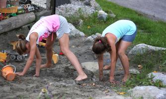Two girls are playing in the sand and digging with their hands. Around them are toys and a green meadow.
