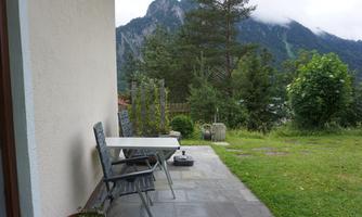 A quiet terrace with a table and chairs, surrounded by green grass and trees. In the background, mountains can be seen with light clouds.