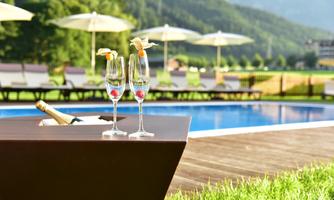Two champagne glasses with fruit are standing next to a pool in the midst of a green landscape. In the background, there are sun umbrellas and mountains visible.