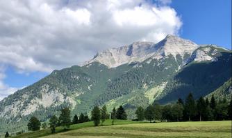 An impressive mountain landscape with green meadows and trees in the foreground. The sky is partially cloudy and the mountains are majestically visible in the background.