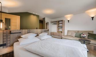 A modern bedroom with a large bed and a cozy sitting area. The walls are in soft colors, and there is harmonious lighting.