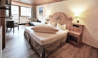 A cozy hotel room with a large bed and wooden details. There is a seating area with a table and plenty of daylight.