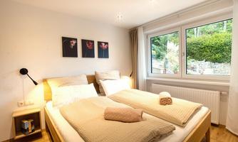 A cozy bedroom with two beds and a large window. The walls are decorated with beautiful pictures and the floor is made of wood.