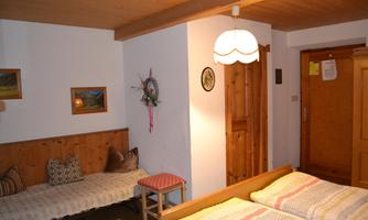 A cozy room with two beds and wooden furniture. Pictures hang on the wall, and there is a small table as well as a lamp on the ceiling.