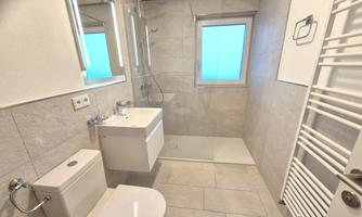 A modern bathroom with a shower and a sink. It is bright and designed in a minimalist style, with large tiles and a window.