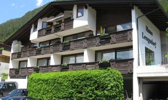 A typical alpine building with wooden balustrades and a well-kept green area. The sky is blue and the surroundings are mountainous.