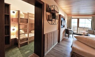 A modern hotel room with wooden-paneled walls and a cozy atmosphere. In the background, a separate children's room with a bunk bed and plenty of daylight can be seen.