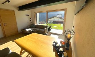A cozy living room with a dining table, sofa, and large windows that let in daylight. Outside, there is a beautiful view of the greenery and the mountains.