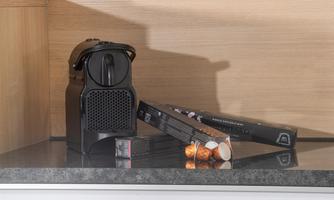 A black coffee machine sits on a granite surface. Next to it are coffee capsules and a black package.
