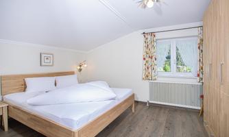 A bright bedroom with a large double bed and wooden floor. The windows are adorned with colorful curtains and offer a view outside.