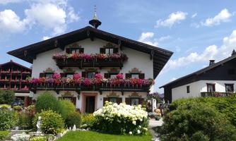 A traditional alpine house with flower balconies and a large garden. The sky is blue and some clouds are visible.