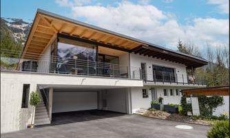A modern house with large windows and an elegant wooden roof. The outdoor area is well-kept and surrounded by greenery.