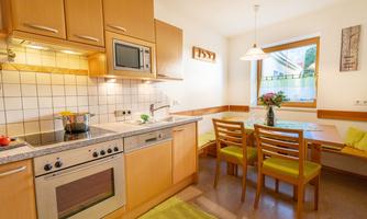 A modern kitchen with light wooden furniture and a cooking area. The dining area has a table with chairs and a large window front that provides plenty of daylight.