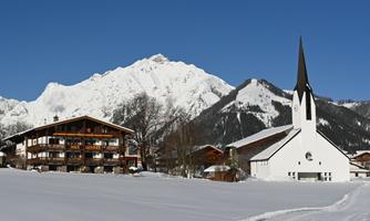 A picturesque winter landscape with snow-covered mountains. In the foreground stands a traditional wooden house and a white church.