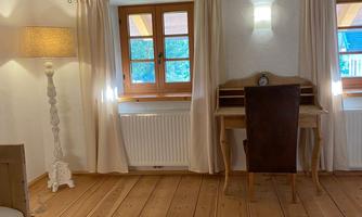 An inviting room with wooden floors and large windows. There is a desk, a lamp, and bright curtains.