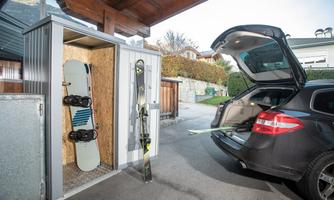 A car with an open trunk is parked next to a shed. Inside the shed, a snowboard and skis are stored.