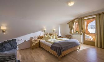 A cozy room with a large bed and natural wooden flooring. Large windows provide a view outside and let in plenty of daylight.