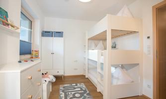 A bright children's room with a bunk bed and a play carpet. There is a wardrobe against the wall and some toys are on a shelf.