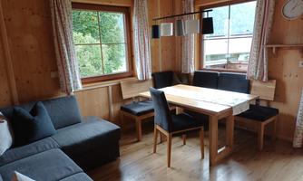 A cozy dining area in a wooden-clad cabin with a large table and comfortable seating. Daylight streams through the windows, offering a beautiful view.