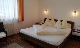 A cozy bedroom with a large bathroom bed and decorative pillows. The room has bright windows and a warm wooden floor.
