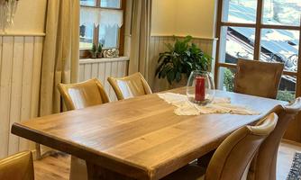 A cozy dining area with a large wooden table and beige chairs. On the table, there is a vase with a red candle, and a green plant is visible in the background.