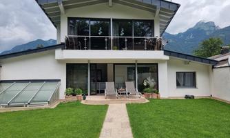 A modern house with large windows and a terrace. The garden is designed with green grass and plants, and mountains can be seen in the background.