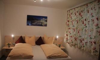 A cozy bedroom with a double bed and colorful cushions. The window has a floral curtain, and a calming picture hangs on the wall.