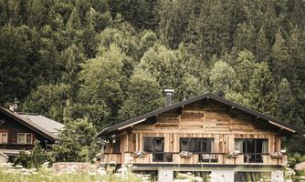 A beautiful wooden house in a green landscape. In the background, tall trees and mountains rise.