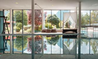 An elegant indoor pool with large windows overlooking a beautiful garden. The surface of the water reflects the surroundings, creating a relaxing atmosphere.