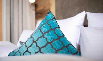 A stylish turquoise pillow with a geometric pattern is lying on a bed. In the background, white pillows and curtains are visible.